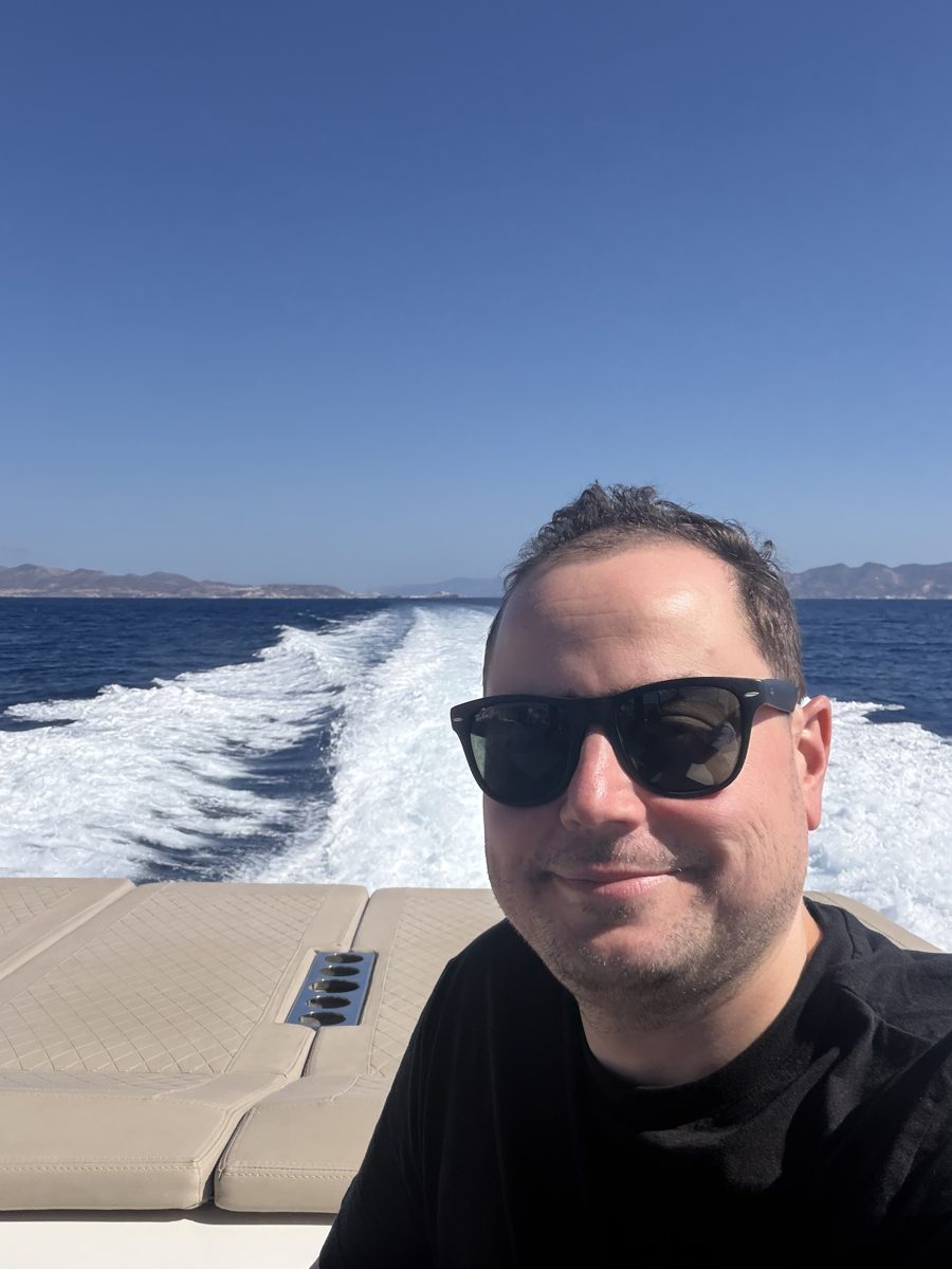 Michael smiling on the back of a boat, with its wake and distant islands behind him